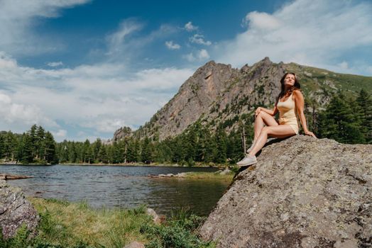 Tourist girl enjoys the magical view of the lake, coniferous forest and magical view sitting on big stone on the shore of a turquoise lake in the mountains. Hiking in the Natural Park. Black lake.