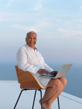 relaxed white hair senior man looking sunset from luxury balclony home and working on laptop computer