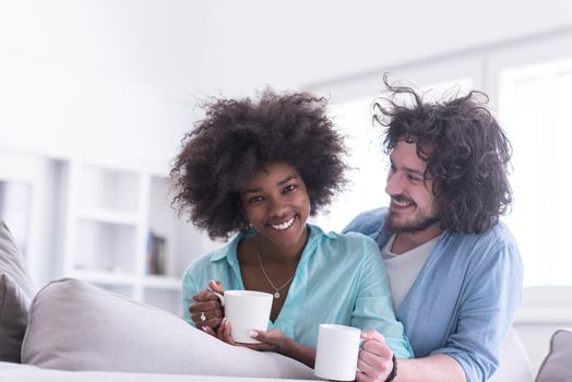 young multiethnic couple sitting on sofa at home drinking coffe, talking, smiling.