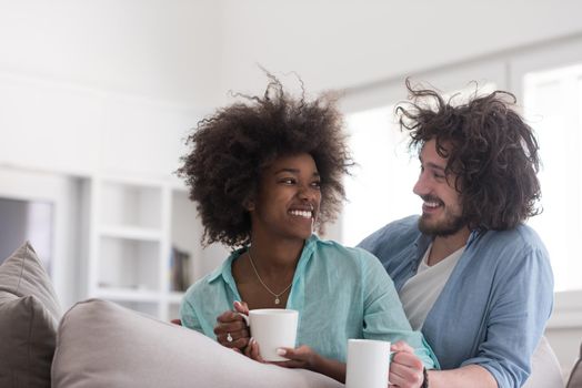 young multiethnic couple sitting on sofa at home drinking coffe, talking, smiling.