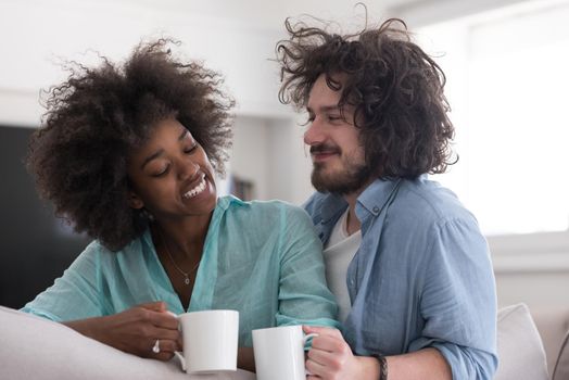 young multiethnic couple sitting on sofa at home drinking coffe, talking, smiling.