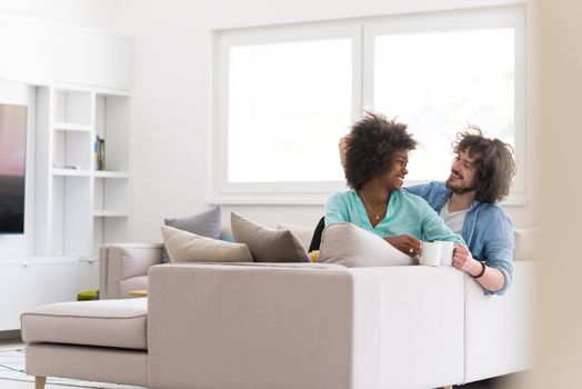 young multiethnic couple sitting on sofa at home drinking coffe, talking, smiling.