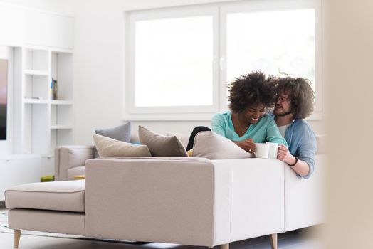 young multiethnic couple sitting on sofa at home drinking coffe, talking, smiling.