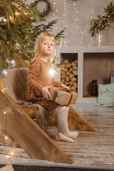 A little blonde girl is sitting on a wooden staircase in a Scandinavian interior decorated in a New Year's style. A child holds a Christmas gift in a craft package.