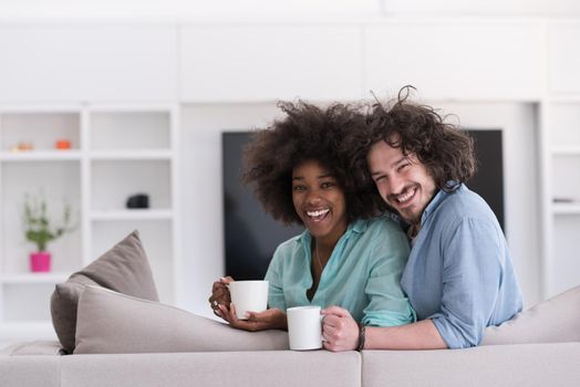 young multiethnic couple sitting on sofa at home drinking coffe, talking, smiling.