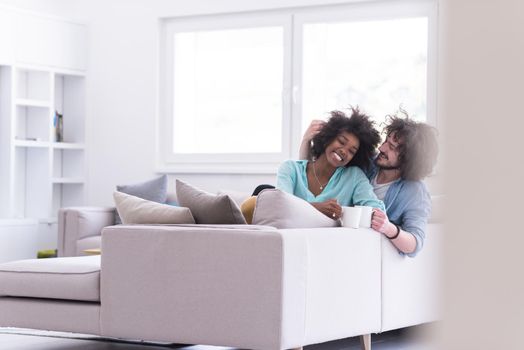 young multiethnic couple sitting on sofa at home drinking coffe, talking, smiling.