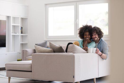 young multiethnic couple sitting on sofa at home drinking coffe, talking, smiling.