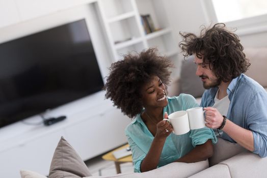 young multiethnic couple sitting on sofa at home drinking coffe, talking, smiling.