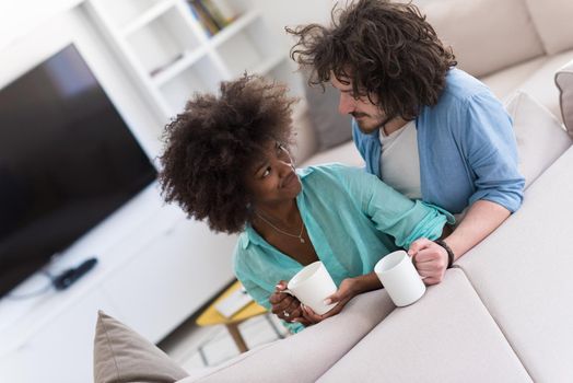 young multiethnic couple sitting on sofa at home drinking coffe, talking, smiling.