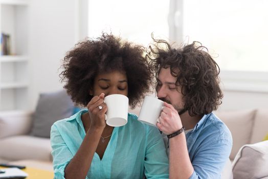 young multiethnic couple sitting on sofa at home drinking coffe, talking, smiling.