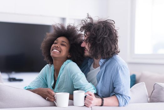 young multiethnic couple sitting on sofa at home drinking coffe, talking, smiling.