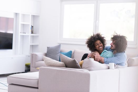 young multiethnic couple sitting on sofa at home drinking coffe, talking, smiling.