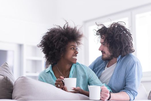 young multiethnic couple sitting on sofa at home drinking coffe, talking, smiling.