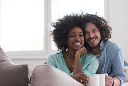 young multiethnic couple sitting on sofa at home drinking coffe, talking, smiling.