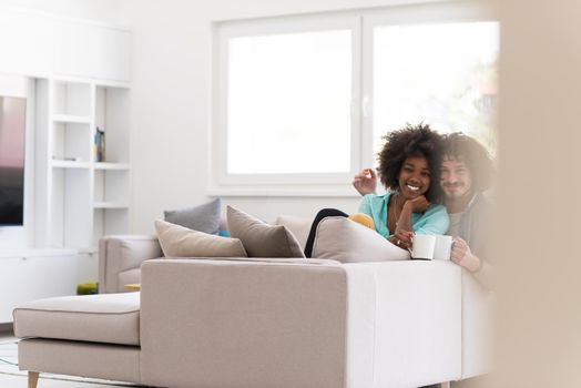 young multiethnic couple sitting on sofa at home drinking coffe, talking, smiling.