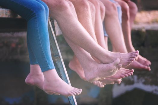 group of  people sitting at wooden bridge over the river with a focus on hanging legs