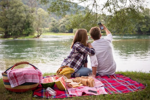 Couple in love taking a selfie by mobile phone while enjoying picnic time drink and food in beautiful nature on the river bank