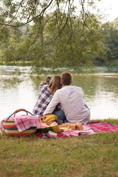 Couple in love taking a selfie by mobile phone while enjoying picnic time drink and food in beautiful nature on the river bank