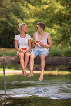 couple in love enjoying watermelon while sitting on the wooden bridge over the river in beautiful nature