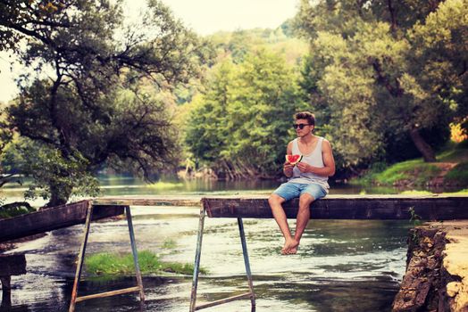 young man enjoying watermelon while sitting on the wooden bridge over the river in beautiful nature
