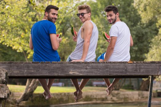 group of young men enjoying watermelon while sitting on the wooden bridge over the river in beautiful nature