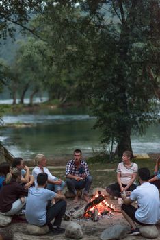 a group of happy young friends relaxing and enjoying  summer evening around campfire on the river bank