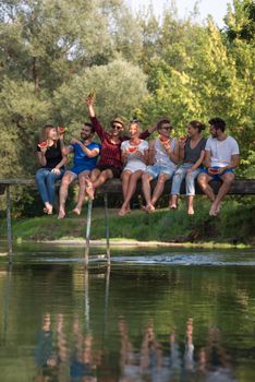 group of young friends enjoying watermelon while sitting on the wooden bridge over the river in beautiful nature