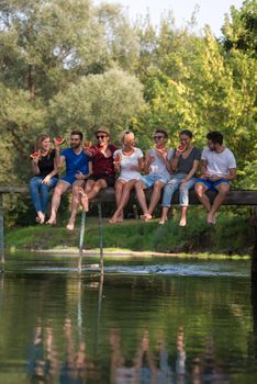 group of young friends enjoying watermelon while sitting on the wooden bridge over the river in beautiful nature