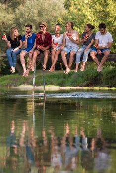 group of young friends enjoying watermelon while sitting on the wooden bridge over the river in beautiful nature