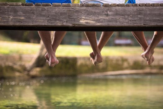 group of  people sitting at wooden bridge over the river with a focus on hanging legs