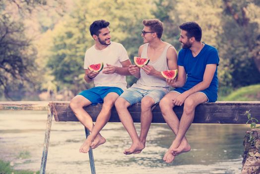 group of young men enjoying watermelon while sitting on the wooden bridge over the river in beautiful nature