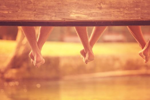 group of  people sitting at wooden bridge over the river with a focus on hanging legs