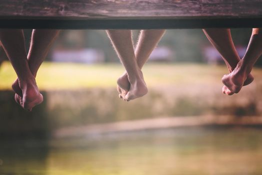 group of  people sitting at wooden bridge over the river with a focus on hanging legs