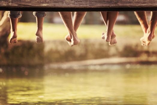 group of  people sitting at wooden bridge over the river with a focus on hanging legs