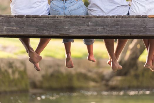 group of  people sitting at wooden bridge over the river with a focus on hanging legs