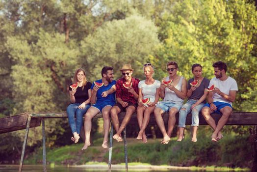 group of young friends enjoying watermelon while sitting on the wooden bridge over the river in beautiful nature