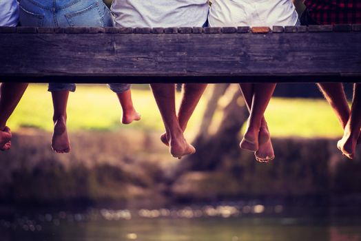 group of  people sitting at wooden bridge over the river with a focus on hanging legs