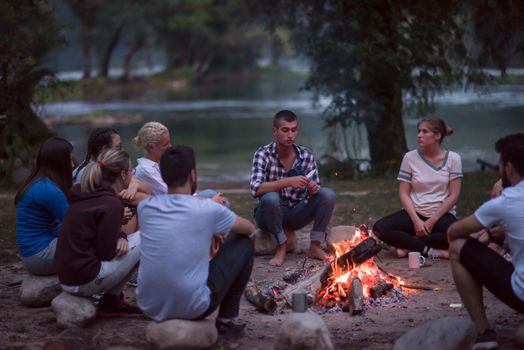 a group of happy young friends relaxing and enjoying  summer evening around campfire on the river bank