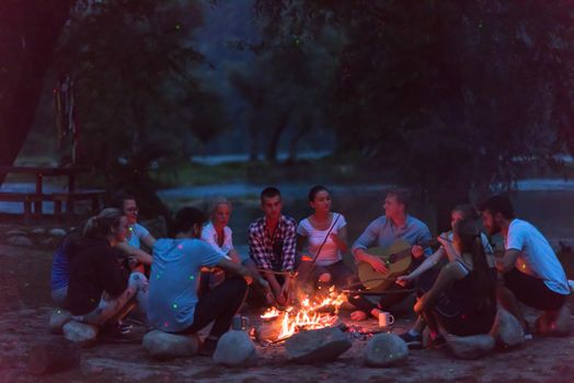 a group of happy young friends relaxing and enjoying  summer evening around campfire on the river bank