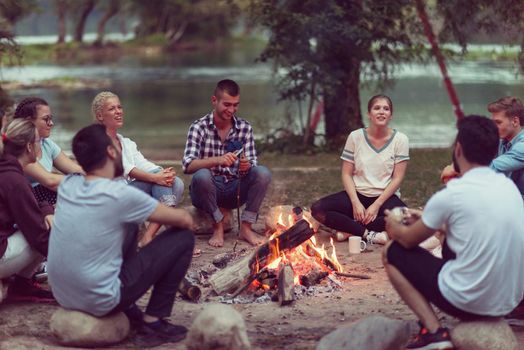 a group of happy young friends relaxing and enjoying  summer evening around campfire on the river bank