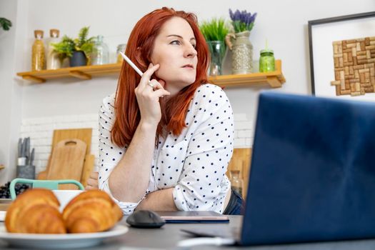 Smiling red haired woman sitting at the table in her dining room drawing on a tablet computer with a stylus, close up