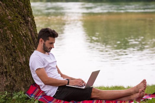 A young freelancer using a laptop computer while working in beautiful nature under the tree on the bank of the river