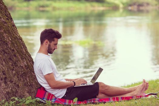 A young freelancer using a laptop computer while working in beautiful nature under the tree on the bank of the river