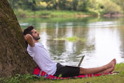 A young freelancer using a laptop computer while working in beautiful nature under the tree on the bank of the river