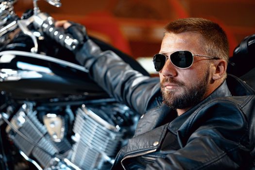 Cool man biker in sunglasses sitting near his motocycle in dark building
