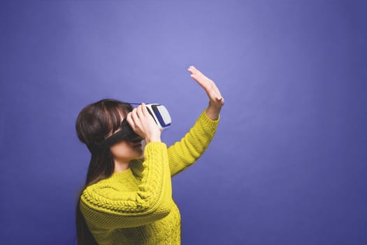A woman in a yellow sweater wearing virtual reality glasses headset, vr box. Demonstrating the colors of 2022 - Very Peri. Changing colors of the year concept. Illuminating and very pari.