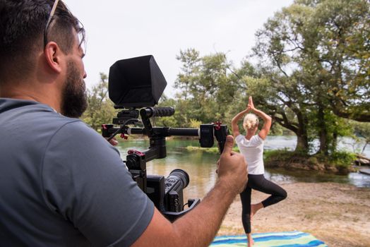 young videographer with gimball video slr rocording while healthy woman doing yoga exercise in the beautiful nature on the bank of the river