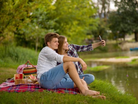 Couple in love taking a selfie by mobile phone while enjoying picnic time drink and food in beautiful nature on the river bank