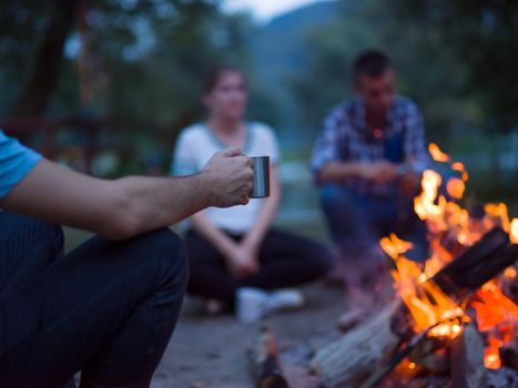 a group of happy young friends relaxing and enjoying  summer evening around campfire on the river bank