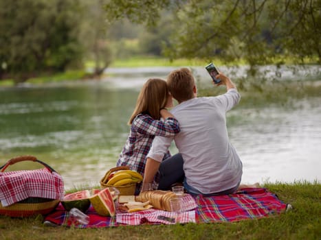 Couple in love taking a selfie by mobile phone while enjoying picnic time drink and food in beautiful nature on the river bank